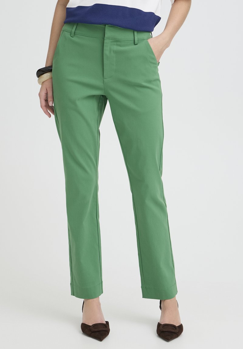 Pantalon vert sur mesure avec une texture lisse et une coupe slim. Doté de passants de ceinture et d'un design à plat à l'avant. Associé à des chaussures marron à talons.
