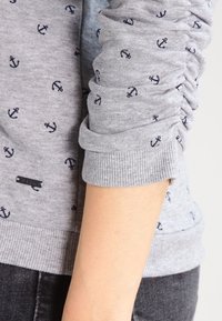 Sudadera gris con patrón de anclas en azul marino, detalle de manga fruncida, puños acanalados y una pequeña etiqueta negra en el dobladillo.