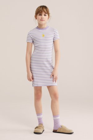 Junges Mädchen steht und trägt ein kurzärmeliges Kleid mit lila-weißen Streifen und einem kleinen Fruchtlogo, lila Socken und beige Slip-on-Schuhe.