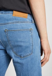 Jeans de mezclilla de color azul claro con un bolsillo trasero visible, costuras amarillas y una etiqueta de cuero marrón con texto en relieve en la cinturilla.