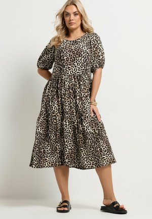 Kobieta stojąca, ubrana w sukienkę midi z falbanami w panterkę, z bufkami na rękawach, oraz czarne sandały ze złotym klamrą.