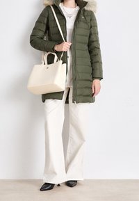 Olivgrüner Daunenmantel mit einer mit Fell gefütterten Kapuze, cremefarbenes Handtasche und weiße, ausgestellte Hosen; schwarze spitze Schuhe runden das Outfit ab.
