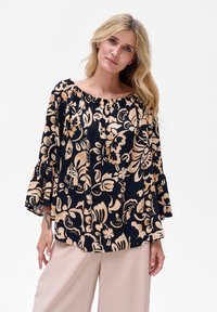 Kwiatowa bluzka na jedno ramię w kolorze granatowym i beżowym. Posiada dzwonkowe rękawy oraz marszczony dekolt. Wykonana z lekkiego materiału o gładkiej fakturze.