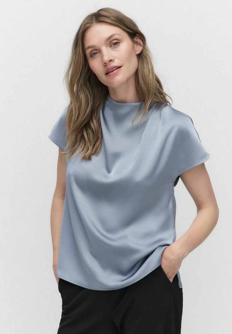 Lyst blå, kortærmet top med draperet design, glat tekstur og høj hals. Bæres med sorte bukser.