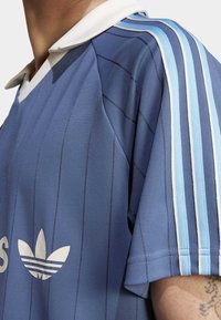 Blå atletisk tröja med vertikala ränder, vit krage, tre vita och ljusblå axelränder samt silverlogotyp framtill.