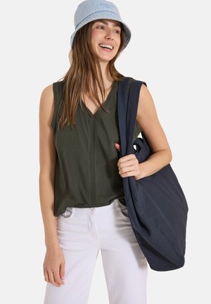 Ung kvinde iført en blå bucket hat og olivengrøn ærmeløs top, der holder en stor mørkeblå tote-taske over skulderen, smilende og kigger til siden.