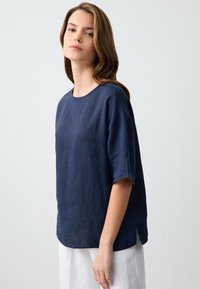Top in lino blu navy con maniche corte, scollatura rotonda e spacchi laterali. Tessuto morbido, vestibilità rilassata e orlo posteriore leggermente più lungo.