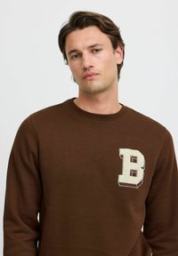 Brun genser i myk stoff, rund hals, med en teksturert hvit bokstav "B" på venstre bryst. Enkel, avslappet design.