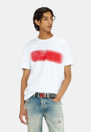 Giovane uomo che indossa una maglietta bianca con un logo rosso sfocato, jeans blu usurati e una cintura grigia con fibbia rossa, mano in tasca.