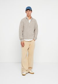 Nuori mies, jolla on päällään sininen lakki, beige neulepaita valkoisen paidan päällä, väljä vaalea housupari ja valkoiset tennarit, seisoo tasaisella taustalla.