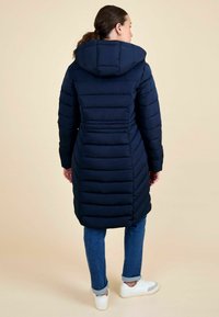 Cămașă puffer bleumarin cu glugă, cu un design matlasat, mâneci lungi și spate strâns. Purtată peste blugi albaștri și adidași albi.
