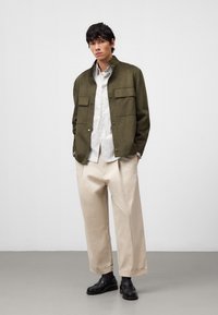 Giacca in suede verde oliva con due tasche frontali, abbinata a una camicia bianca a righe e pantaloni larghi beige chiaro. Scarpe nere completano il look.