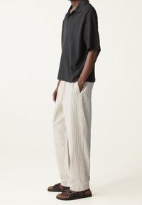 Camisa negra de manga corta con tejido texturizado, pantalones anchos de rayas crema y sandalias oscuras. Los pantalones cuentan con bolsillos laterales y un corte holgado.