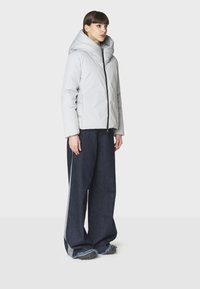 Giacca puffa grigio chiaro con colletto alto e chiusura a zip, abbinata a jeans in denim scuri dalla vestibilità ampia con dettagli laterali.