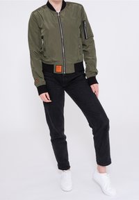 Oliwkowa bomberka z zamkiem z przodu, ściągaczami u mankietów oraz naszywką na talii. Posiada kieszeń na suwak na rękawie.