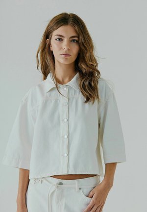 White cropped button-up shirt with short sleeves, made of denim fabric. Features a pointed collar and front placket with circular buttons.
