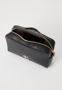 Zwarte lederen cosmetische tas met een gouden ritssluiting, afgeronde hoeken en een goudkleurige logo-detail aan de voorkant. Het interieur is bekleed met een grijze stof.