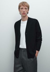 Svart cardigan med avslappnad passform, med V-ringning, knappstängning, ribbade muddar, i kombination med en vit t-shirt och randiga byxor.
