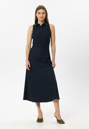 Woman stands wearing a sleeveless navy blue collared midi dress with a tied waist and olive green heeled mules on a plain white background.