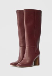 Μπορντό δερμάτινες μπότες μέχρι το γόνατο με λεία υφή, τετράγωνη μύτη και χοντρό τακούνι για σταθερότητα. Ελάχιστες λεπτομέρειες ραψίματος.