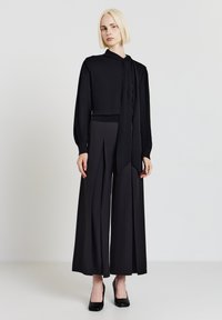 Top nero a maniche lunghe con sciarpa annodata al collo, abbinato a pantaloni neri a vita alta e gamba larga e tacchi neri. Texture liscia.