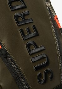 Sac à dos en tissu vert olive doté d'un texte noir brodé audacieux et d'une tirette orange, avec une surface texturée et une forme élégante.