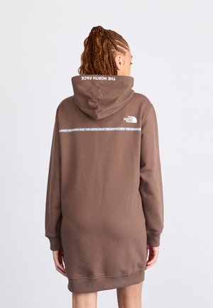 Osoba z zaplecionymi włosami, ubrana w brązową sukienkę z kapturem North Face, zwrócona tyłem, prezentująca logo marki oraz napis na plecach i kapturze.