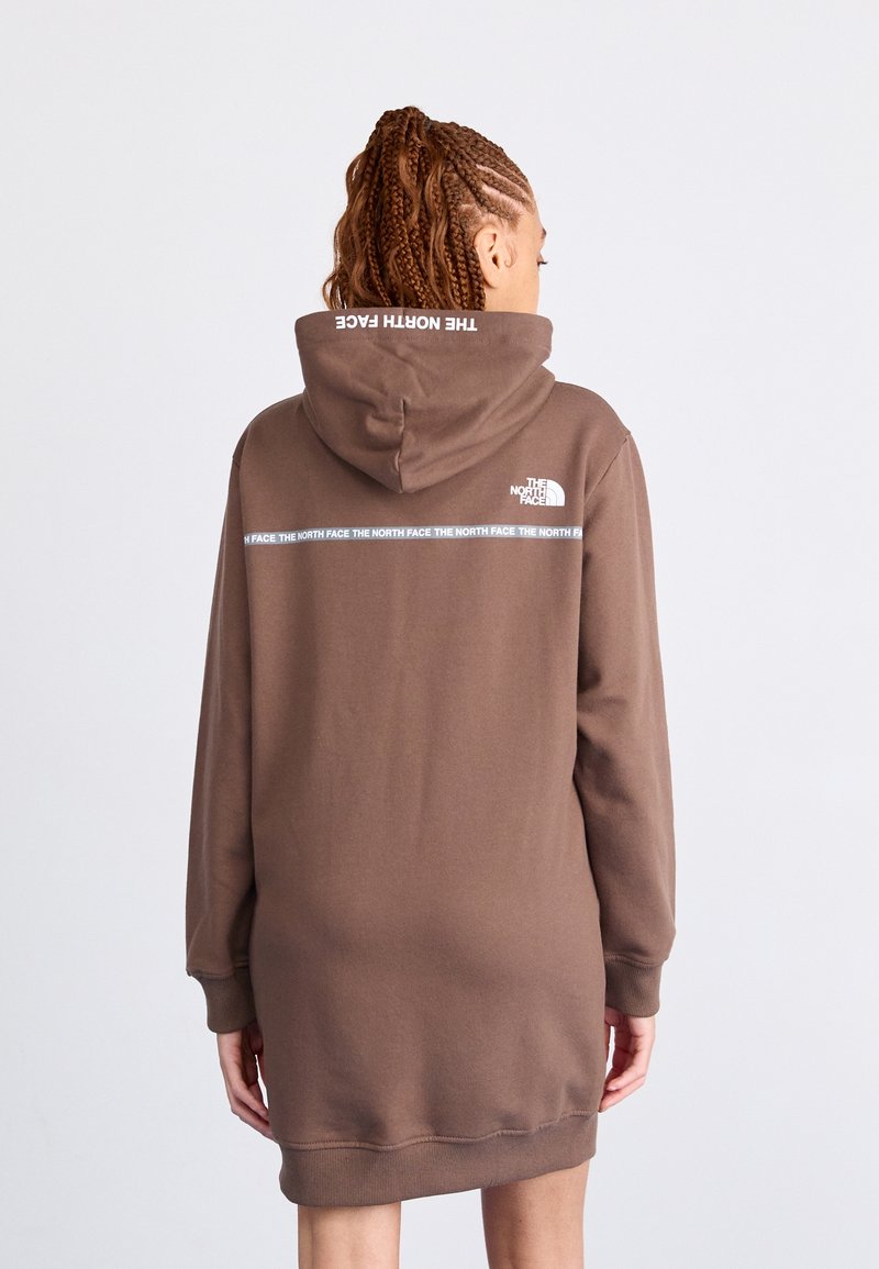 Personne aux cheveux tressés portant une robe sweat à capuche marron de la marque North Face, dos tourné, montrant le logo de la marque et le texte au dos et sur la capuche.