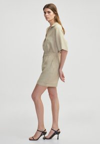 Robe chemise en lin beige clair avec des manches courtes, une taille élastique et une longueur au genou. Associée à des escarpins noirs à lanières.