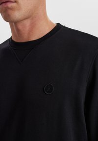 Sort sweatshirt med tekstureret ribkant ved halsen og et lille rundt logo på venstre bryst. Glat stof, afslappet pasform.