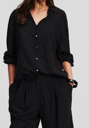 Woman wearing loose black button-up shirt with rolled-up sleeves, black high-waist pleated pants, and a black bracelet, hands in pockets.