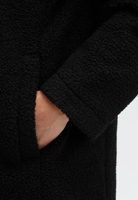 Black fleece coat detail featuring a textured fabric, showing a hand in a side pocket. The material appears soft and fluffy.