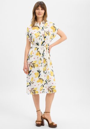 Femme portant une robe midi blanche avec imprimé citron jaune et floral, manches courtes, taille ceinturée et sandales marron à talons hauts.
