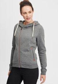 Siva zip-up dukserica s kapuljačom na vezanje, kontrastni smeđi patentni zatvarač i trokutasti detalji na džepovima. Mekani materijal s glatkom teksturom.