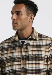 Geruite button-up shirt in beige, bruin en marineblauw. Heeft twee borstzakken, een opgerold kraag en een zachte, gestructureerde stof.