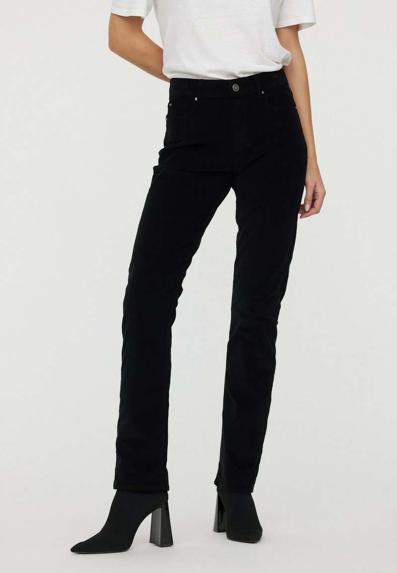 Pantalons en velours côtelé noirs à jambe droite avec une taille haute, présentant des poches avant et une fermeture à bouton. Portés avec des bottines à talons noires.