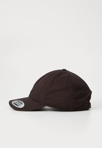 Gorra de tela marrón oscuro ajustable con visera curvada, diseño de seis paneles y costuras visibles. Presenta una etiqueta circular en un lado.