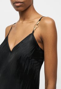 Η γυναίκα φοράει μαύρο σατέν φόρεμα με διακριτικό σχέδιο φύλλων και λεπτές τιράντες που συνδέονται με χρυσούς κρίκους σε σχήμα φίδι στον ώμο.