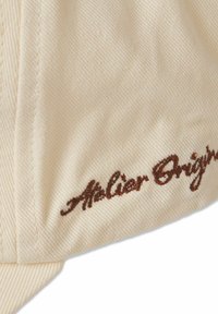 Cream-colored cap with a textured fabric; features brown embroidered "Atelier Origins" text and curved brim.