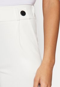 Bílé kalhoty s plochým pasem, zdobené výrazným černým knoflíkem a bočními sklady, hladkou texturou tkaniny a přiléhavým střihem.