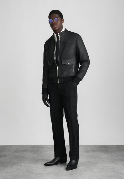 Zwarte leren bomberjack met ritssluiting, twee voorzakken en geribbelde manchetten, gecombineerd met getextureerde zwarte broek en zwarte leren laarzen.