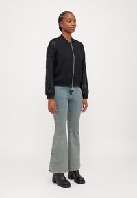 Veste bomber noire avec des manches en sequins, fermeture éclair et poignets côtelés, associée à un jean flare bleu clair et des bottes noires à talons épais.
