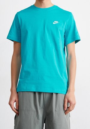 Persoon draagt een felblauw Nike T-shirt en grijze cargoshorts, staat ontspannen met armen langs het lichaam tegen een effen achtergrond.