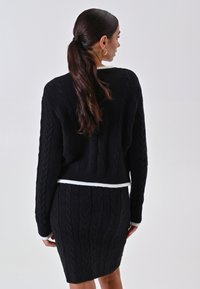Set di maglione e gonna nera in maglia, presenta un design a treccia, polsini e orlo a costine, con una fascia bianca in evidenza in vita.