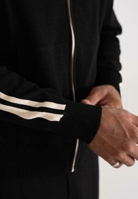 Nærbillede af en person iført en sort zip-up sweater med hvide striber på ærmerne, der justerer ærmekanten med begge hænder.