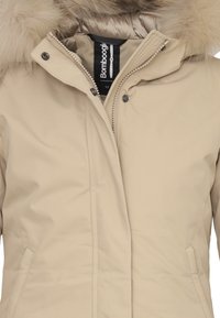 Giacca isolante beige con cappuccio in pelliccia sintetica rimovibile, chiusura con zip e tasche laterali; texture liscia e design minimalista.