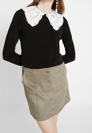 Vrouw die een zwart shirt met lange mouwen draagt met een witte kanten kraag en een beige rok met knopen aan de voorkant, poserend met haar handen achter haar rug.