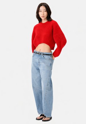 Junge Frau, die einen kurzen roten Pullover, hoch taillierte lockere hellblaue Jeans, schwarzen Gürtel und schwarze offene Sandalen trägt, steht vor einem weißen Hintergrund.