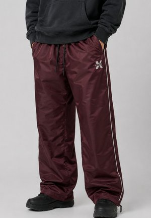 Persona che indossa pantaloni della tuta bordeaux larghi con strisce bianche laterali, coulisse nera, scarpe nere e felpa nera con cappuccio con le mani nelle tasche.