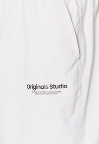 Bijele kratke hlače od tkanine s džepom, pojasom na vezanje i otisnutim brendiranjem "Originals Studio" u crnoj i srebrnoj boji.